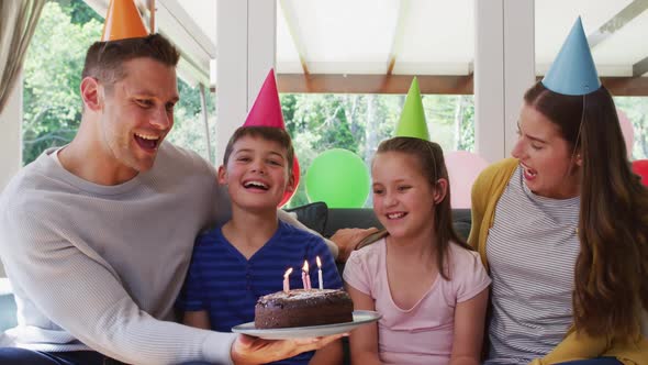 Portrait of caucasian girl in party hat blowing candles on birthday cake while family smiling alt