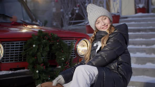 Portrait Smiling Girl on Red Car with Christmas Tree Branch Decoration Background in Winter Day alt