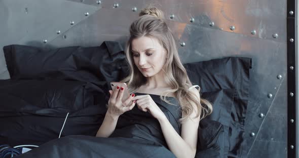 Gorgeous Blondie Checking Her Mobile Phone Sitting in Bed Under Black Blanket