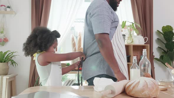 African American dad tying apron on daughter before make bread dough in kitchen at home