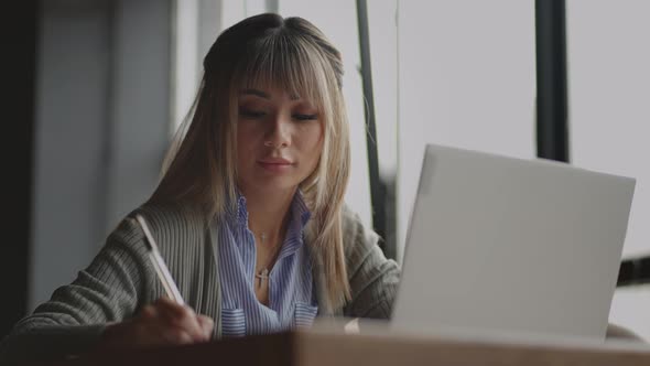 Asian Woman Working on Her Laptop and Writing in His Notebook Sitting at a Table alt