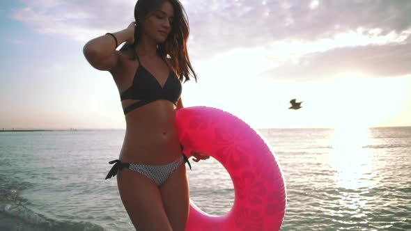 Beautiful Young Woman Walking with Colorful Inflatable Ring at Sea Coast During Sunset Slow Motion alt