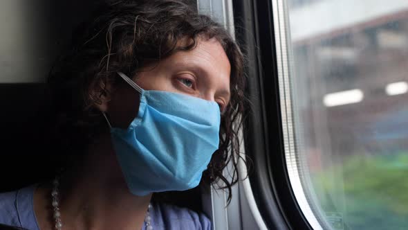A tired middle-aged woman in a protective blue mask looks out the window of a train