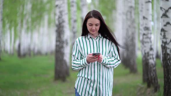 Beautiful Girl with a Smartphone in Her Hands Walks Along a Birch Grove in Spring alt