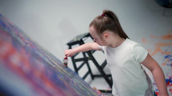 Kid Girl with Down Syndrome Draws with a Brush on a Large Canvas in a White Room Kid Girl with alt