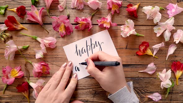 Writing HAPPY ANNIVERSARY on a card near flowers top view alt