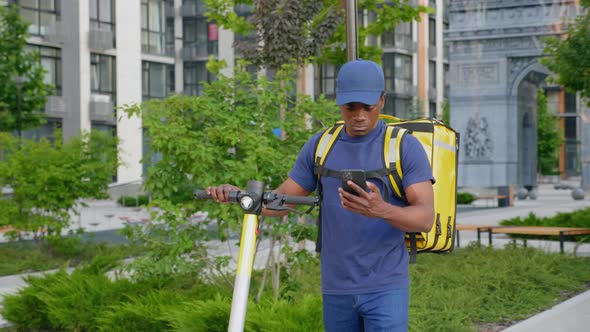 Afroamerican Man Courier Walks with Electric Scooter Uses Smartphone Navigate alt