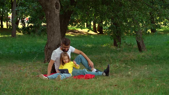 Father and Child Sit on Lawn in Summer Park with Tablet alt