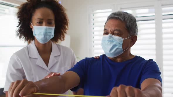 Mixed race female physiotherapist wearing mask helping senior exercise using exercise band alt