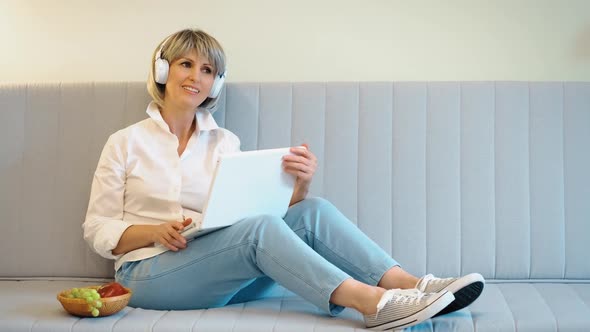 An Elderly Beautiful Woman with Headphones and a Laptop in Her Hands Listens to Music and Sings