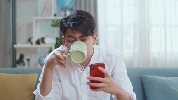 Happy Asian Man Drinking Coffee While Using Smartphone In The Living Room alt
