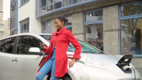 African American Girl Using Smartphone and Leans on Electric Car That Is Charging. Plug Charging an alt