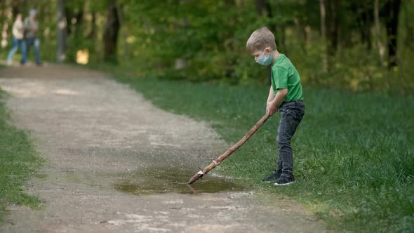 A Small Boy in a Medical Mask Plays with a Stick Near a Puddle in the Park in Spring. The Concept of