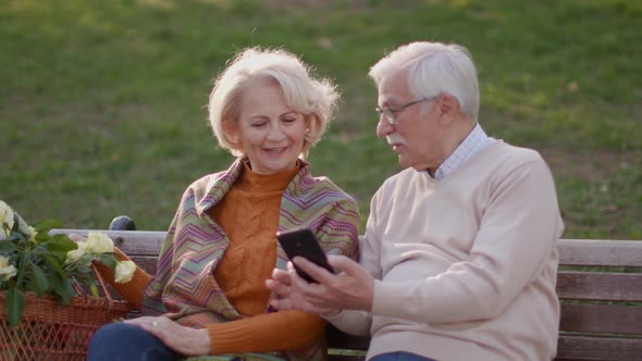 Handsome senior couple sitting on the bench with basket full of flowers and looking at mobile phone