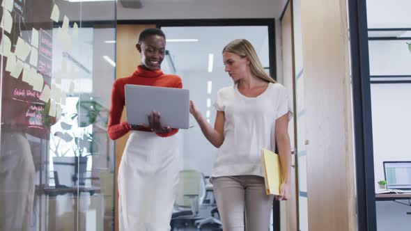 Two diverse female office colleagues with laptop discussing together while walking in office alt