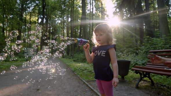 Happy Kid Blows Bubbles From Special Manual Bubble Machine Outdoors in Park alt