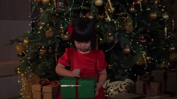 Korean Girl Child is Sitting at the Christmas Tree and Opens Gift Green Box alt