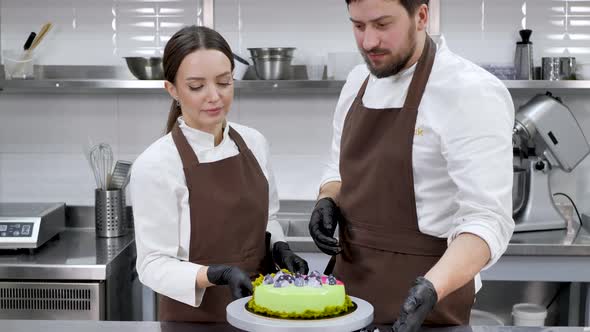 Two Pastry Chefs a Man and a Woman Twist a Cake on a Stand in a Professional Kitchen alt