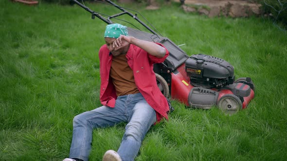 Wide Shot of Tired Perspiring Young Man Drying Forehead with Hand Sitting on Green Backyard with alt