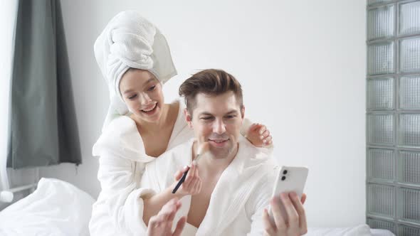 Cheerful Young Caucasian Couple in Bathrobes and Towel Have Fun Woman Doing Makeup on Boyfriend's