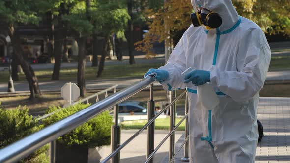 A Virologist in Protective Clothing Disinfects the Railing with an Antiseptic Spray and a Tissue alt