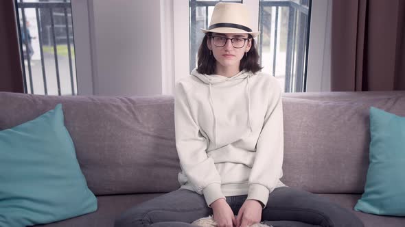 Girl in Glasses and a Hat Sits on the Sofa with Her Legs Crossed Window Background alt