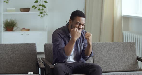 Afro American Guy at Home on Couch Cheering for Beloved Player in Boxing Competition Online alt
