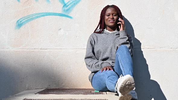 Young Black Woman Sitting Near the Concrete Wall and Speaking on a Phone