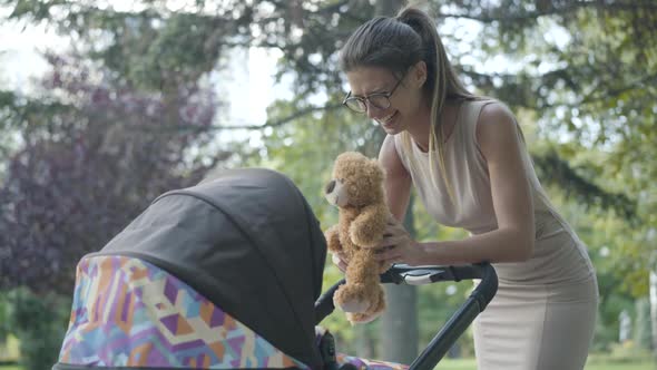 Charming Young Caucasian Woman in Eyeglasses Playing Teddy Bear and Laughing Admiring Infant in Baby alt