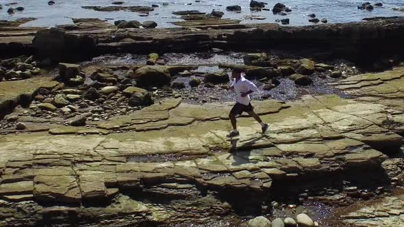 Side view tracking shot of a young man running on a rocky ocean beach shoreline. alt