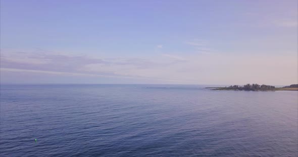 Aerial shot flying over a vast deep blue ocean with a small island beach in the distance and blue cl alt