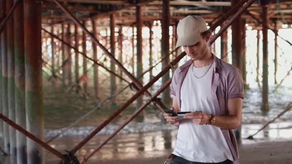 Man standing Under Pier Paying For a Product Using His Smart Phone and Credit Card alt