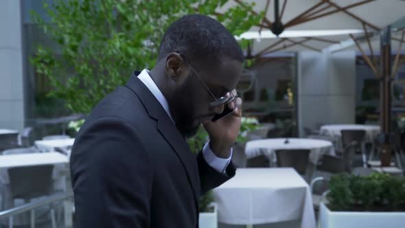 Male in Business Suit Passing by Luxury Open Air Cafe Talking Over Cell Phone alt