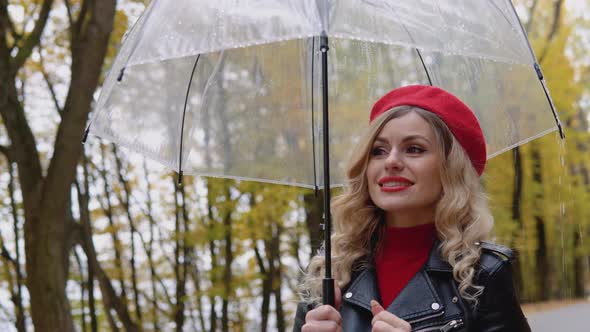 Romantic Blonde in a Red Dress and Biker Jacket with Transparent Umbrella Standing in Park alt