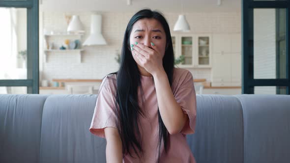 Young Darkhaired with Long Hair Woman of Asian Descent Looks at Camera Sitting on Sofa and Suddenly alt