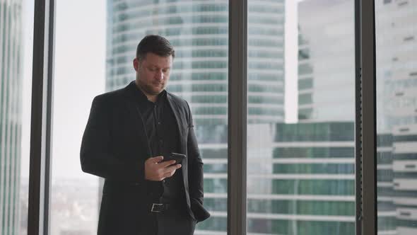 A Businessman Stands with His Back to the Window Against the Backdrop of a Skyscraper alt