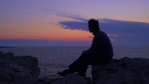 Young Man Waits for the Sunrise at the Sea. Blue hour twilight, gazing at the horizon. Middle Shot alt