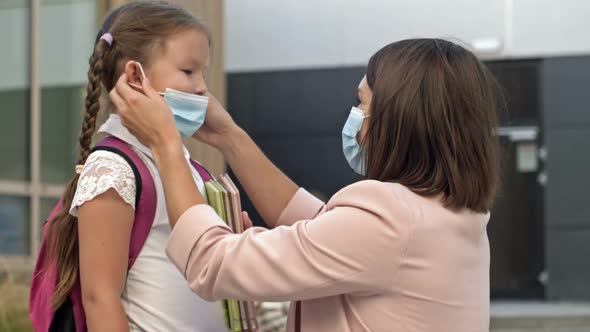 Young Woman Wearing a Protective Mask Puts on Her Daughter's Mask Before Entering the School