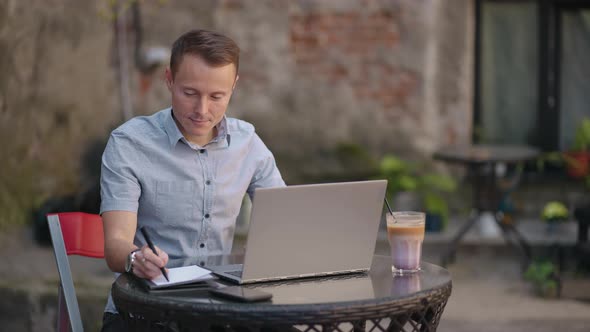 A Man Sitting in a Street Cafe Works Remotely Writing a Pen in a Notebook alt