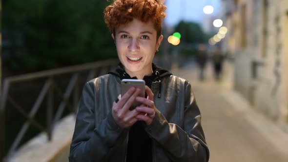 Young caucasian curly redhead woman outdoor in city sunset holding smart phone alt