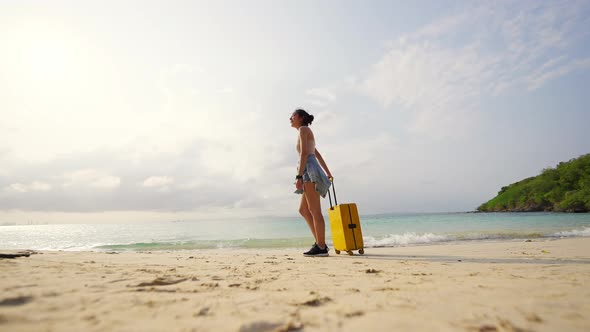 4K Asian woman dragging a suitcase and walking on tropical beach at summer sunset.