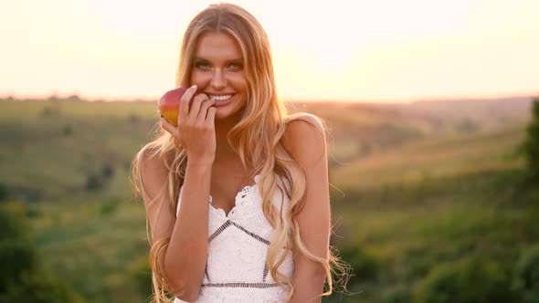 Beautiful sexy blonde girl in white dress posing in a field at sunset with a basket of fruit