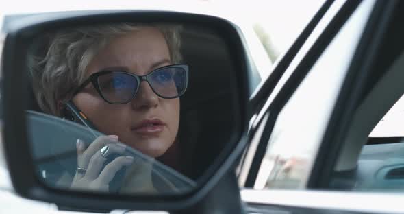 Stylish Woman in Glasses Talking on a Phone While Driving Car View Through Rear Mirror