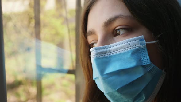 A Sad Girl in Medical Mask Looks Out the Window alt