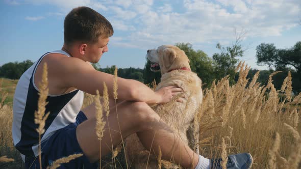Young Man Sits on Grass at Meadow and Strokes His Labrador or Golden Retriever alt