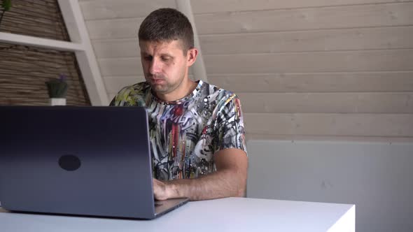 An Adult Man with a Laptop Works at Home at the Table alt