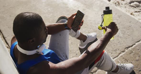 African american man sitting and using smartphone taking break in exercise outdoors alt