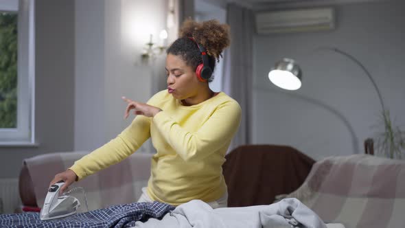 Cheerful African American Woman in Headphones Ironing Laundry and Dancing Having Fun alt