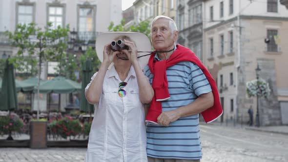 Senior Male and Female Tourists Walking in Town Center and Looking in Binoculars alt