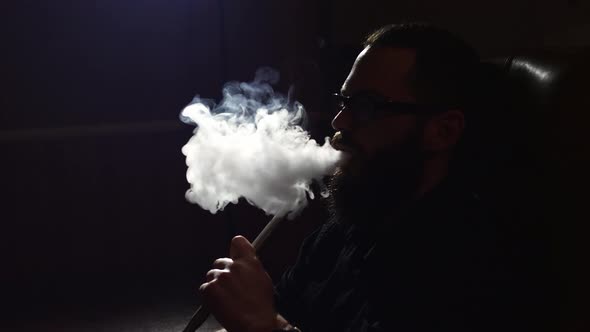 Young Bearded Man in Glasses Smokes a Hookah and Blow Out Smoke Closeup on Black Background in Slow